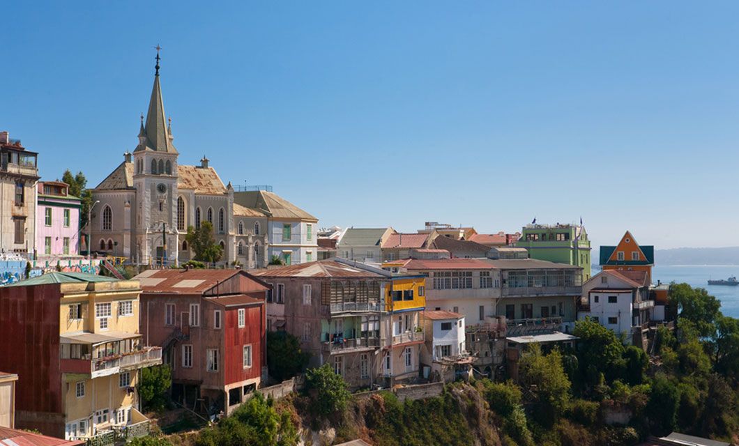 A hillside neighborhood with colorful houses and a large church with a tall steeple under a clear blue sky, overlooking the ocean in Valparaíso, Chile—a scenic stop on the journey from Santiago to Buenos Aires or before a Patagonia cruise.