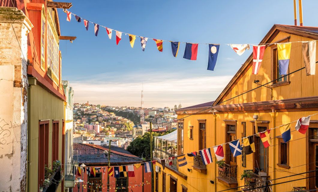 Colorful houses with yellow walls line a narrow street decorated with strings of international flags; the cityscape and distant hills are visible under a clear blue sky, just a short drive from serene lakes and picturesque beaches.
