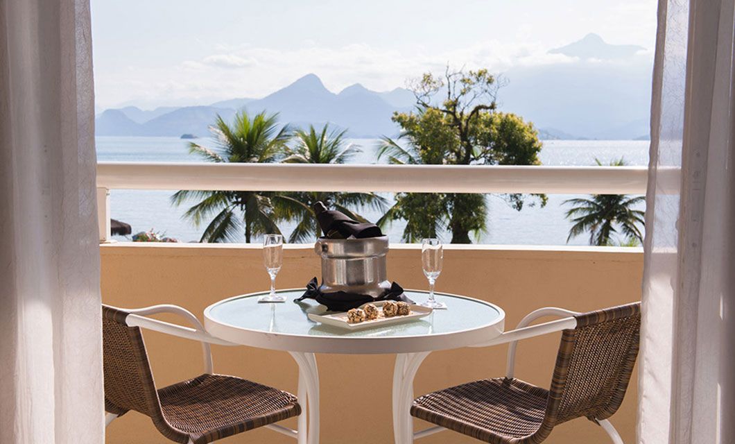A small round table with two wicker chairs on a balcony at Vila Gale Angra dos Reis overlooks the sea and distant mountains. Two glasses, an ice bucket with a bottle, and pastries await, with palm trees and blue water framing this Brazil beach resort view.