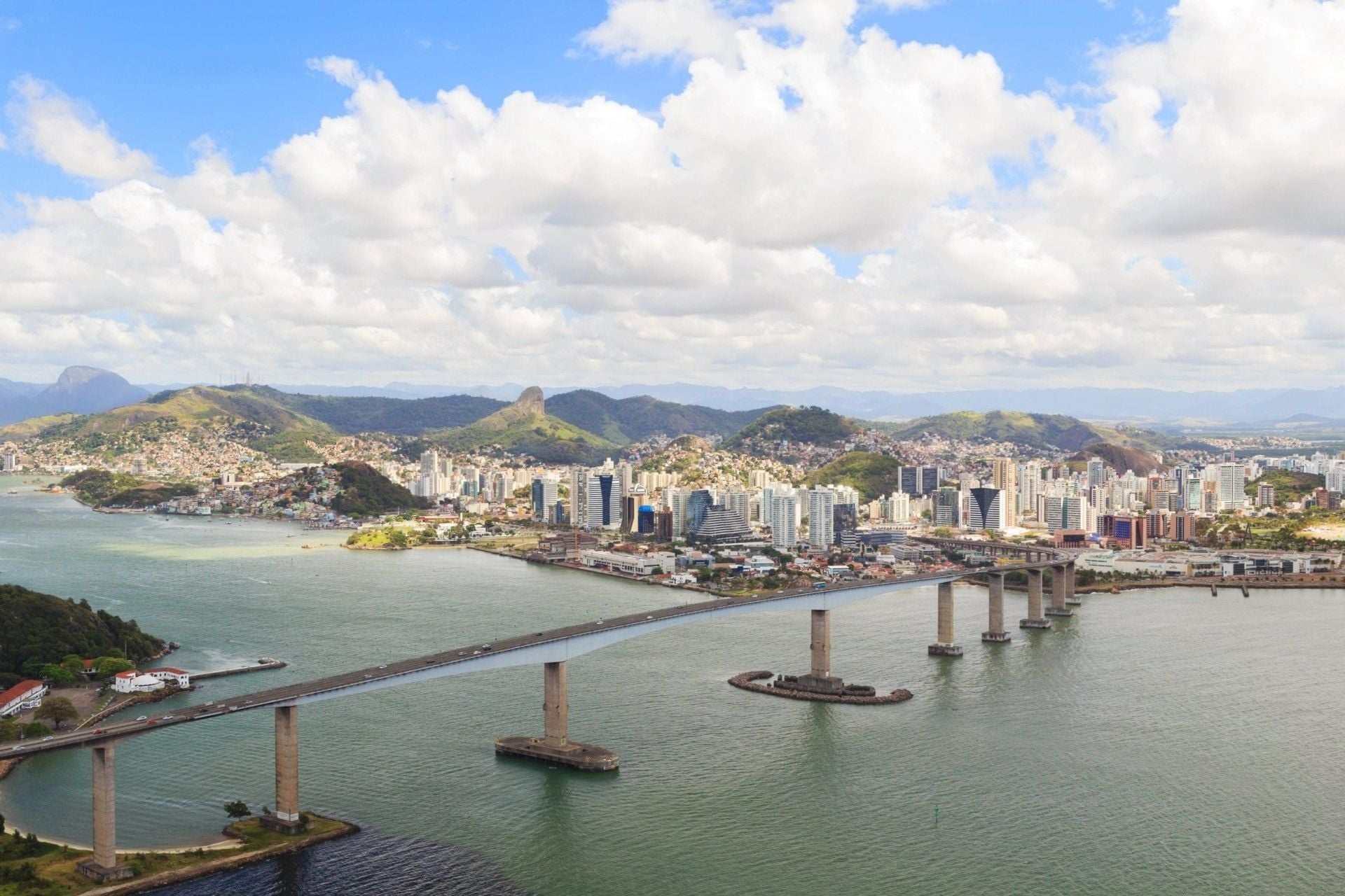 A long bridge spans a wide body of water, leading to a city with tall buildings and green hills in the background, evoking the scenic views often experienced on a Brazil Train Tour under a partly cloudy sky.
