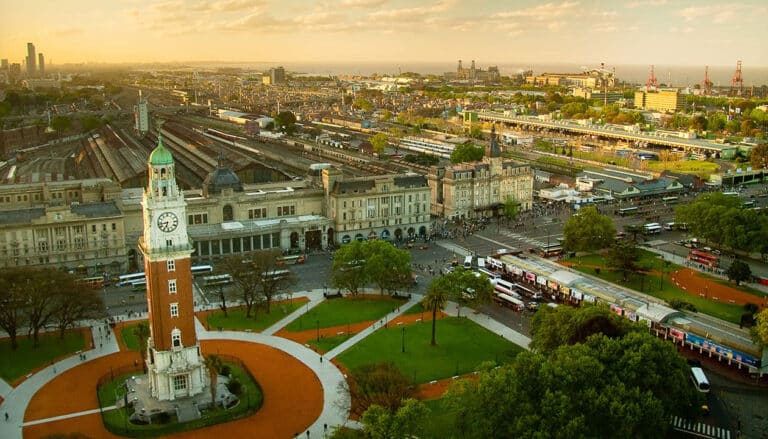Aerial view of a cityscape with a tall clock tower in a park, large historic railway station behind it, green trees, and glimpses of lakes, people, and city buildings extending into the distance under a partly cloudy sky.