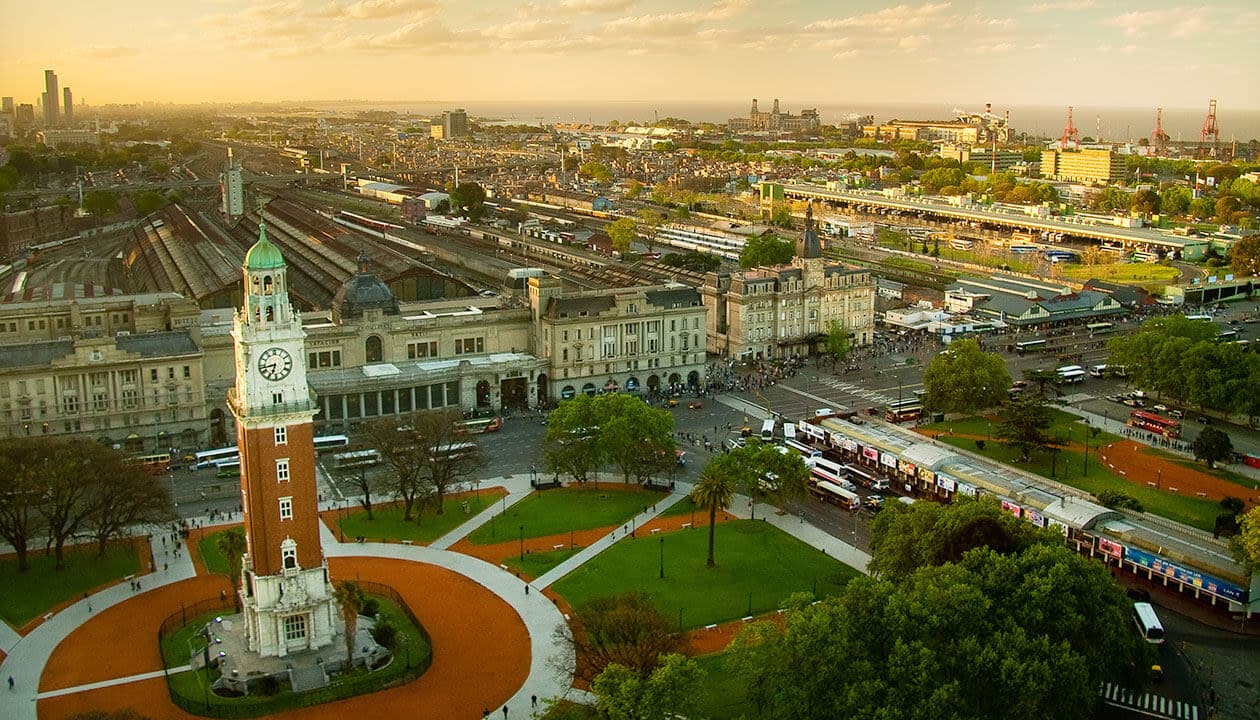 Aerial view of a cityscape with a tall clock tower in a park, large historic railway station behind it, green trees, and glimpses of lakes, people, and city buildings extending into the distance under a partly cloudy sky.