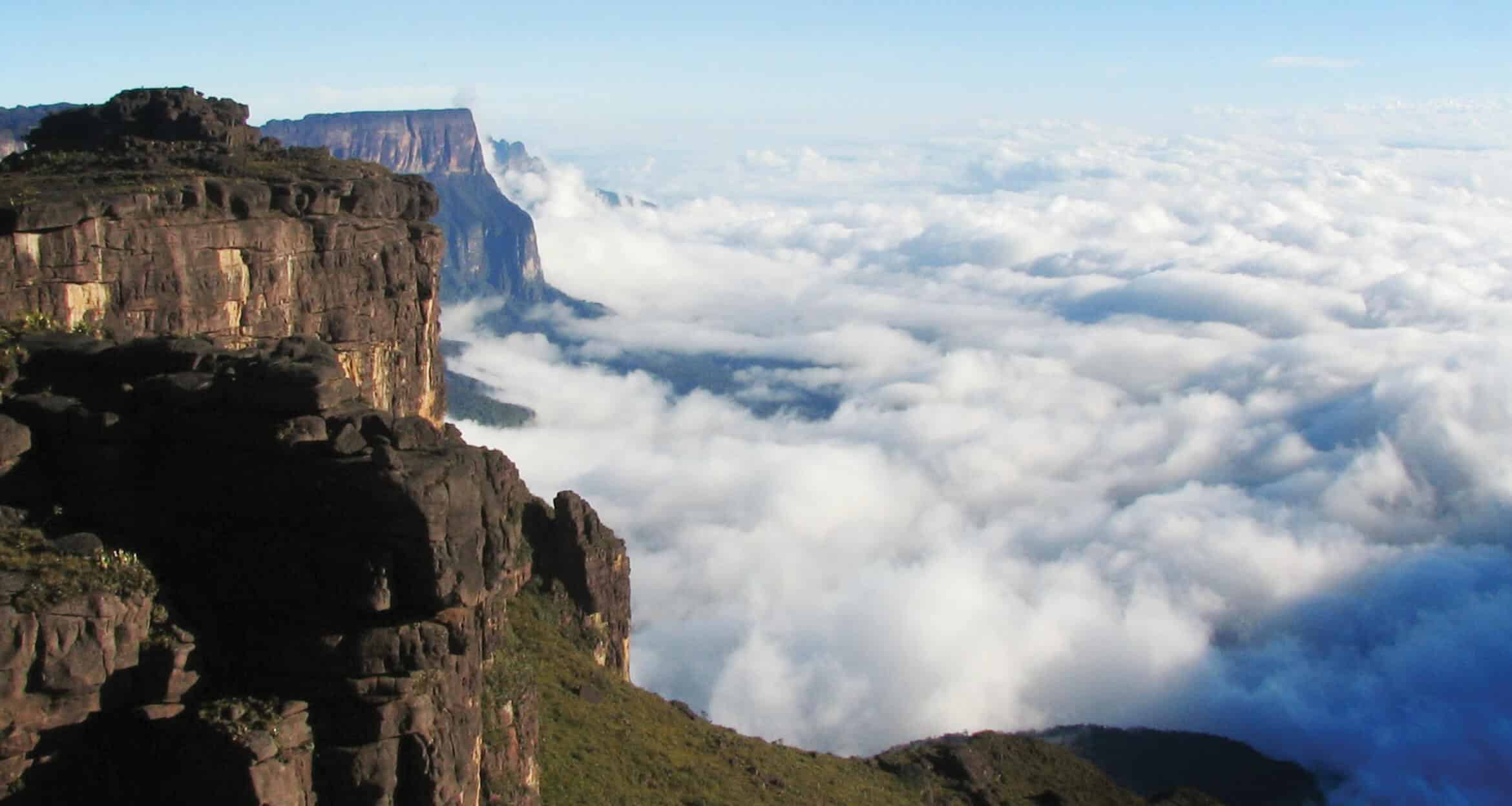 Steep rocky cliffs rise above a blanket of dense white clouds on Mount Roraima, with sunlight illuminating the rugged terrain. In the distance, more plateaus emerge from the sea of clouds under a clear blue sky.