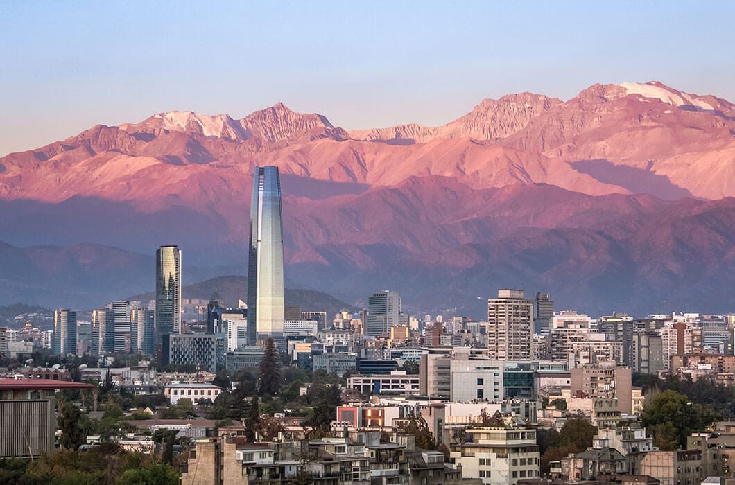 A city skyline with tall modern buildings stands before snow-capped mountains at sunset, the peaks bathed in pink light—a scene that captures the magic of South American Wonders.