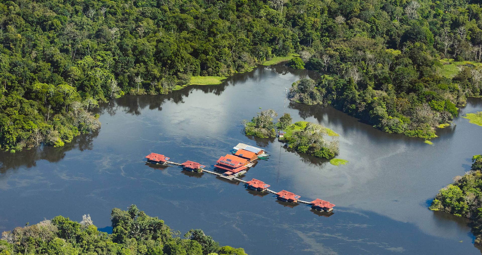 Aerial view of floating lodge buildings with red roofs on a calm river in the Amazon rainforest, surrounded by dense green trees and lush vegetation.