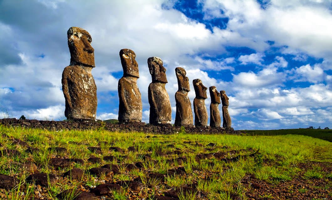 Seven large stone Moai statues stand in a row on grassy terrain under a partly cloudy blue sky on Easter Island, a must-see stop on any tour from Santiago. The statues face away from the camera, surrounded by rocks and patches of green grass.