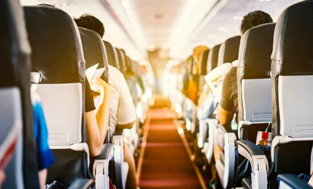 View from the aisle of an airplane cabin, showing passengers seated in rows, reading or relaxing, as sunlight streams through the windows—perhaps dreaming of serene beaches or majestic waterfalls awaiting them at their destination.