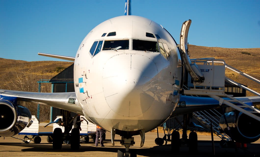 A front view of a large white commercial airplane on the tarmac, its boarding stairs extended, welcomes passengers for a Colombia tour. People and equipment are near the aircraft, set against dry hills under a clear blue sky.