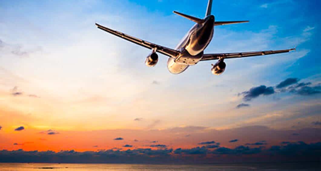 A commercial airplane flies above the ocean with a vibrant sunset sky in the background, reminiscent of the colorful views seen on a Colombia Train Tour from Bogota to Zipaquira, with shades of orange, yellow, and blue scattered among the clouds.