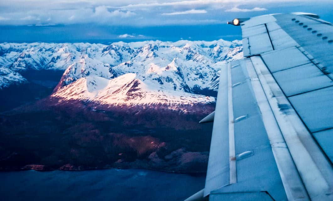 View from an airplane window showing a wing with snow-capped mountains in Patagonia below, sunlight illuminating the peaks above a dark blue body of water.