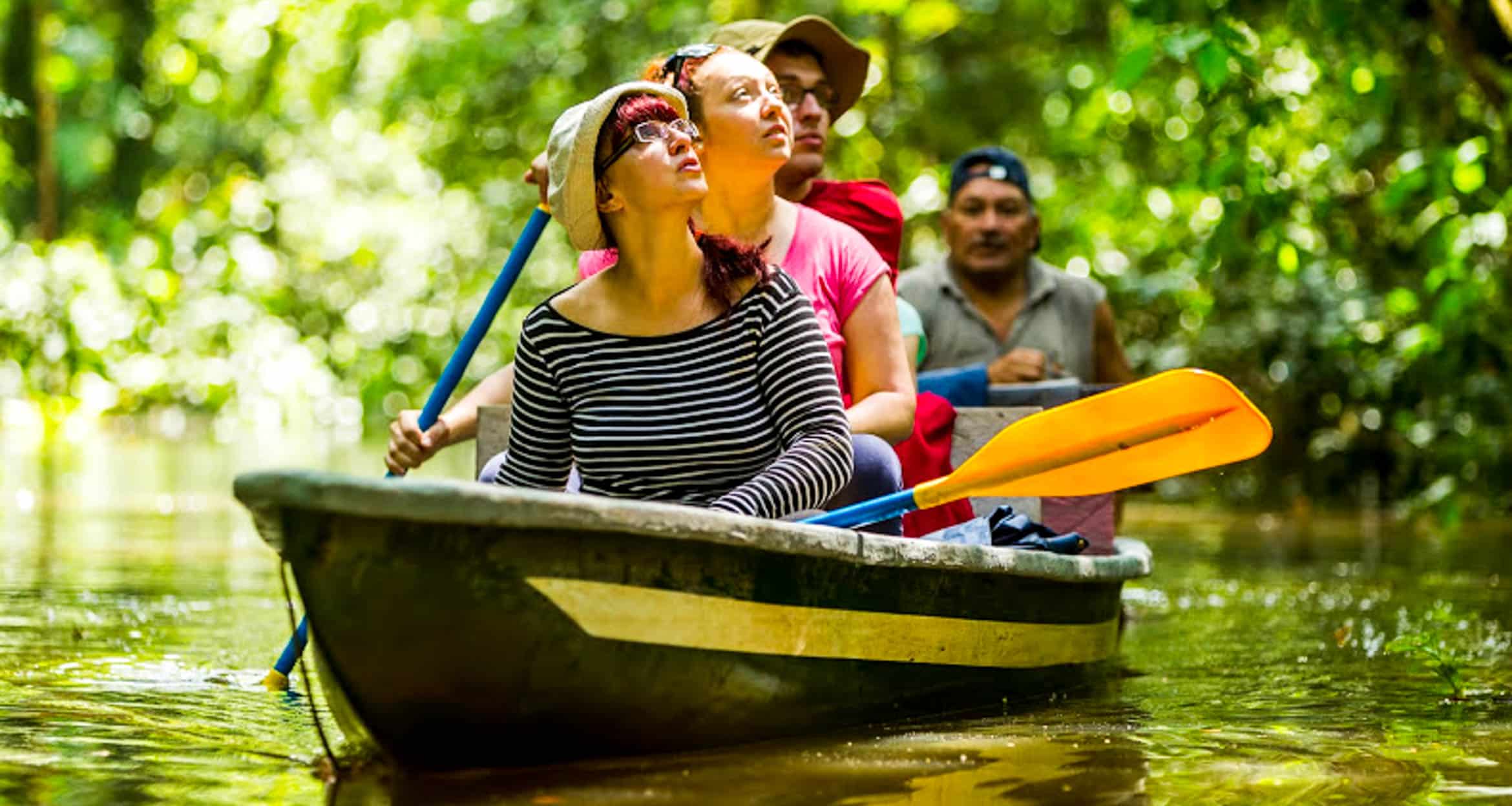 Four people paddle a canoe through a lush Amazon Rainforest waterway, looking up at the towering trees. The calm water and dappled sunlight create a serene atmosphere, making this an unforgettable tour through nature’s wonders.