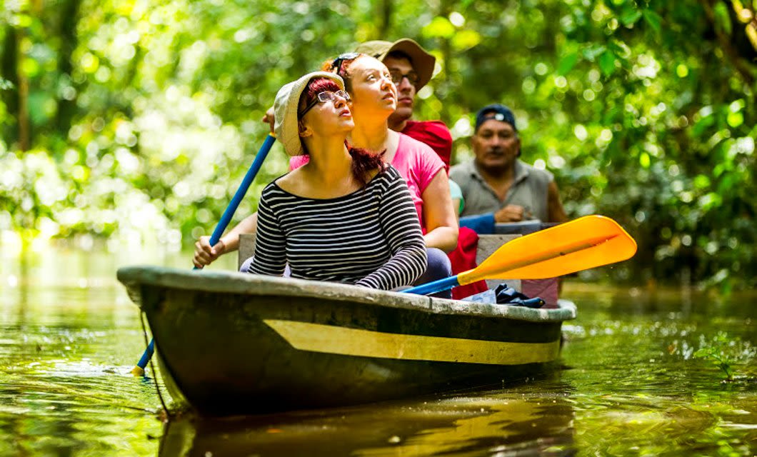 Four people sit in a narrow canoe, paddling through a lush Ecuador Amazon Rainforest waterway. The front passenger looks upward as sunlight filters through dense foliage—an unforgettable scene from an Eco Lodge or Quito tour adventure.