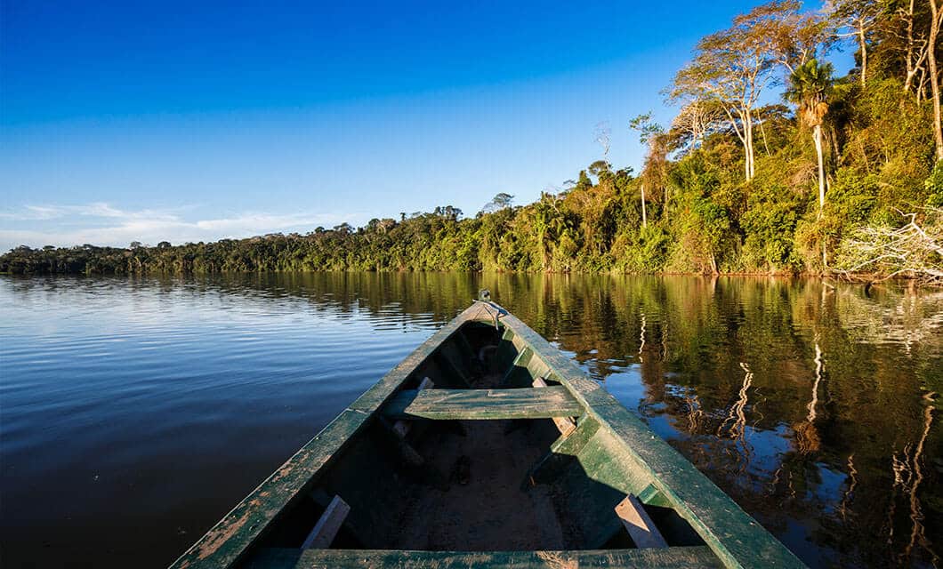 A wooden canoe floats on a calm river during a Remote Amazon Tour, surrounded by lush green forest under a clear blue sky, with dense trees reflected in the water.