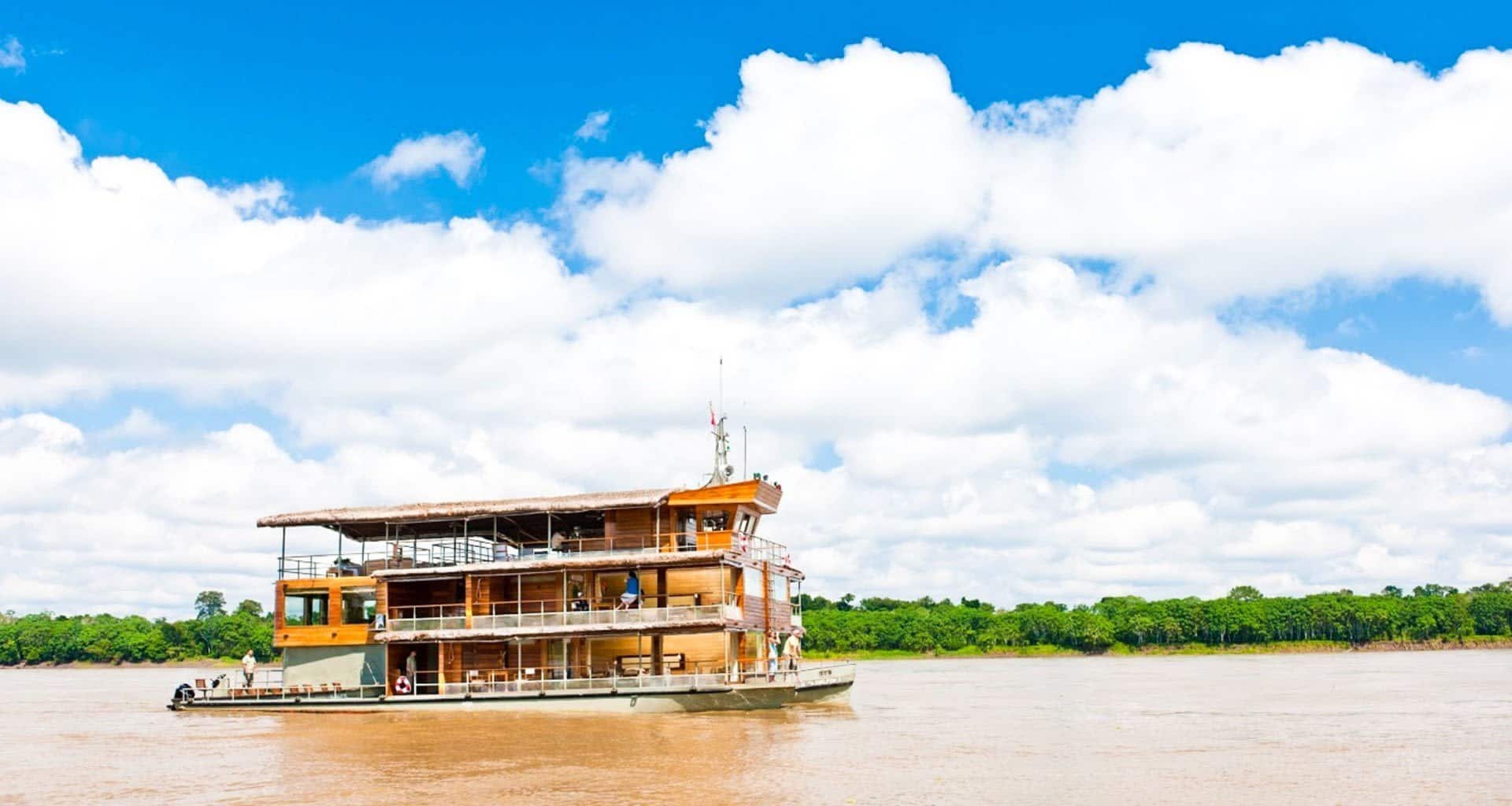 A large wooden riverboat with two decks cruises on a wide, muddy river under a bright blue sky with scattered clouds. Green trees line the distant riverbank, offering a classic scene from an unforgettable Amazon cruise.