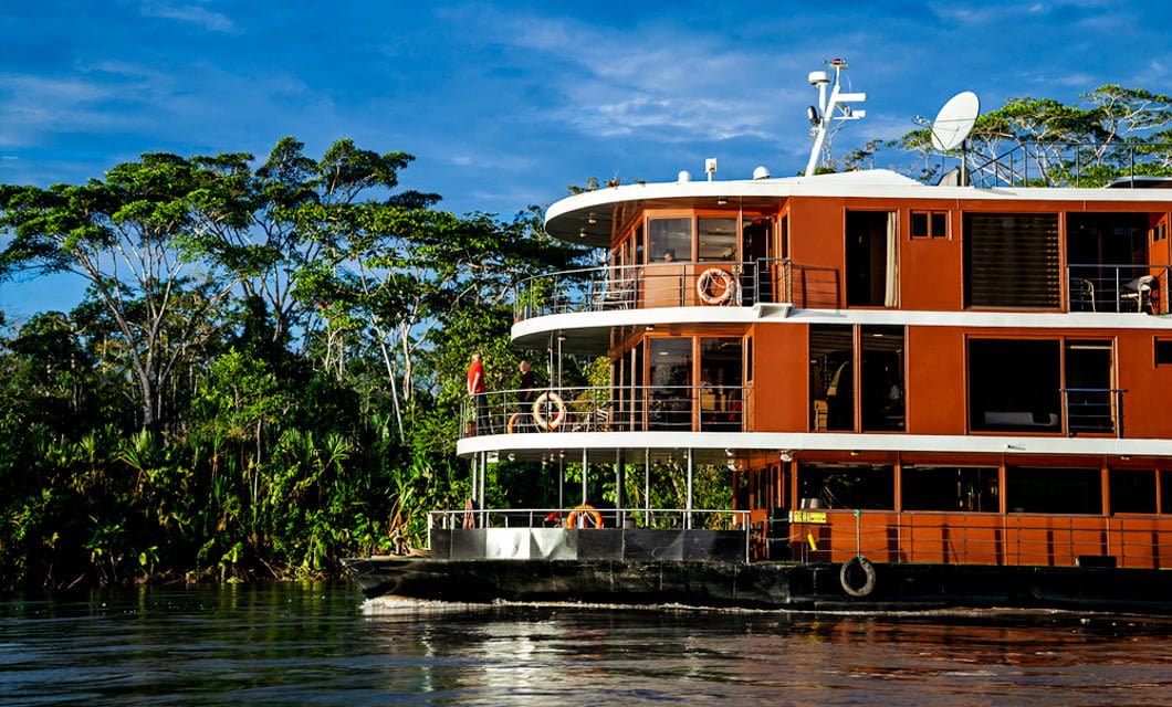 A modern, multi-level riverboat with an orange and white exterior sails the Amazon River, surrounded by lush green trees under a blue sky—an iconic scene from Anakonda Amazon Cruises.