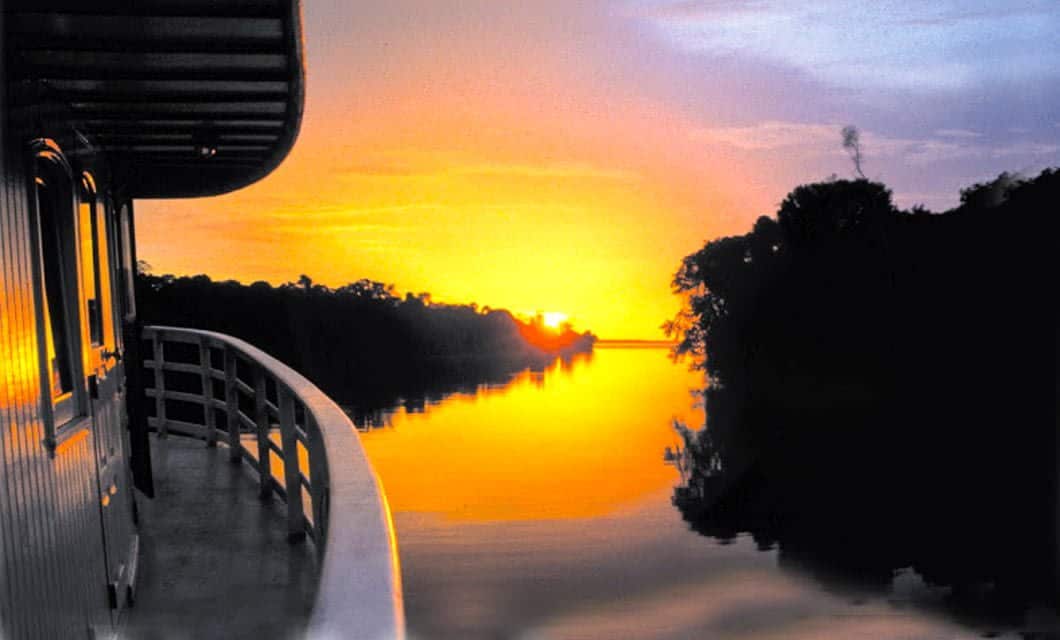 A boat sails on a calm river at sunset during a Brazil Amazon River Cruise, with the sun glowing orange and yellow on the horizon. Trees line the riverbank, and the sky reflects on the water’s surface.