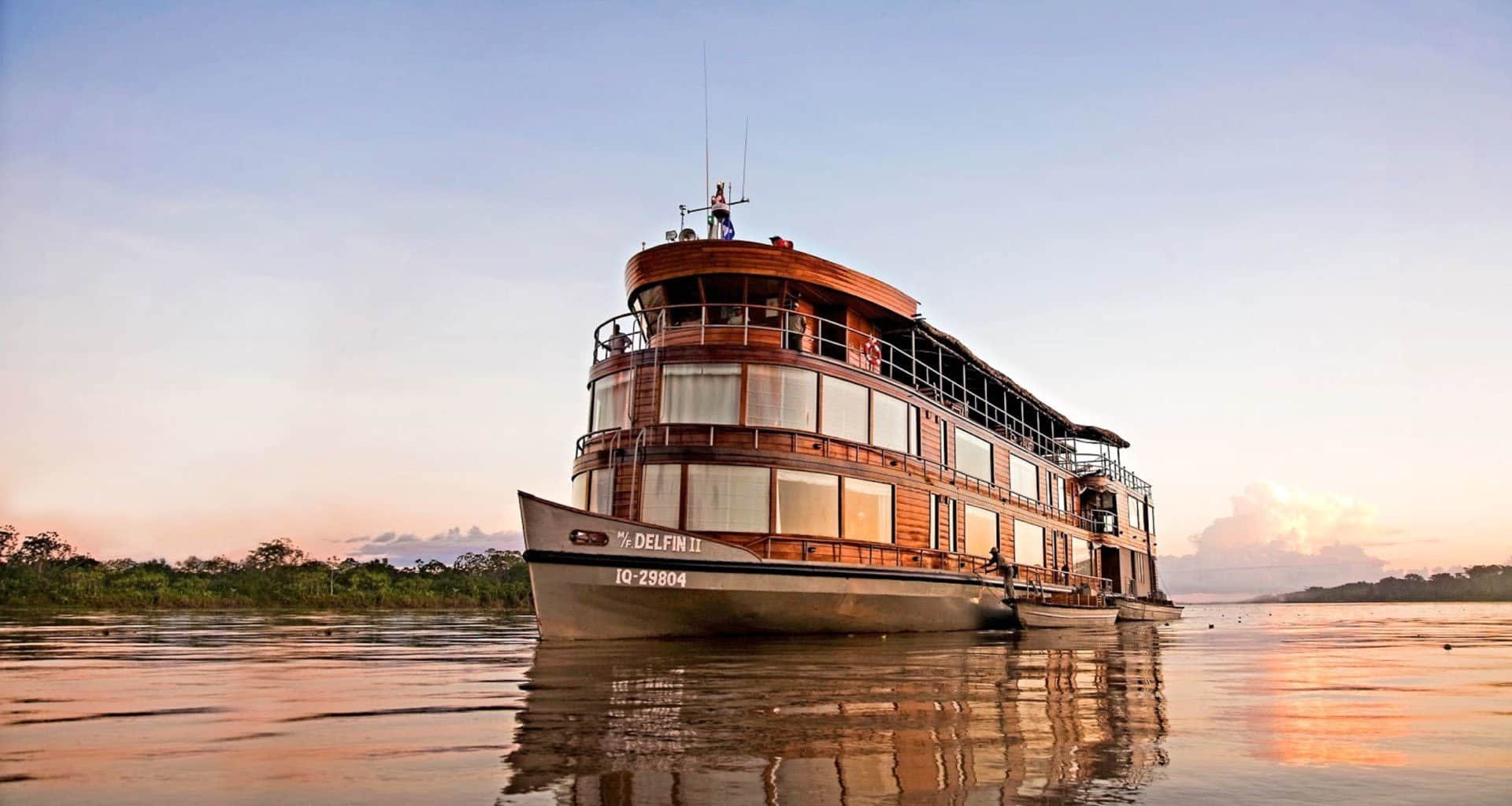 A wooden riverboat with multiple decks, named Delfin I, floats on calm water at sunset, evoking the elegance of an Amazon Delfin II Cruise amid lush green trees and a pastel sky in the Peruvian rainforest.