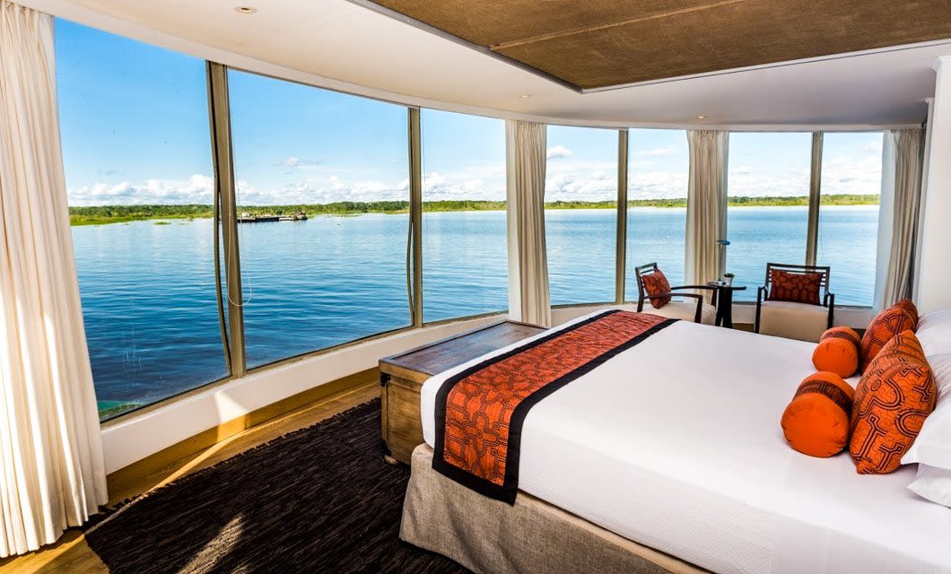 A bright hotel room with a large bed and orange accents, featuring floor-to-ceiling curved windows overlooking a serene lake reminiscent of views from an Amazon luxury cruise. Two chairs and a small table sit near the window.