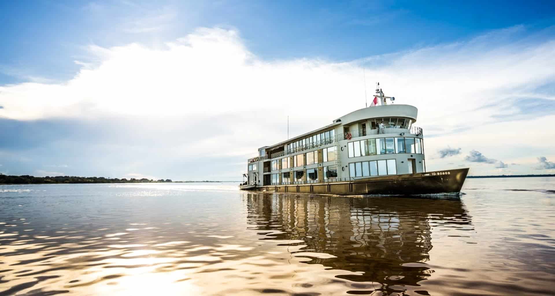A modern Amazon luxury cruise ship sails on calm water under a bright, partly cloudy sky, with sunlight reflecting on the water’s surface. Land is visible in the distance on the left side of the image.