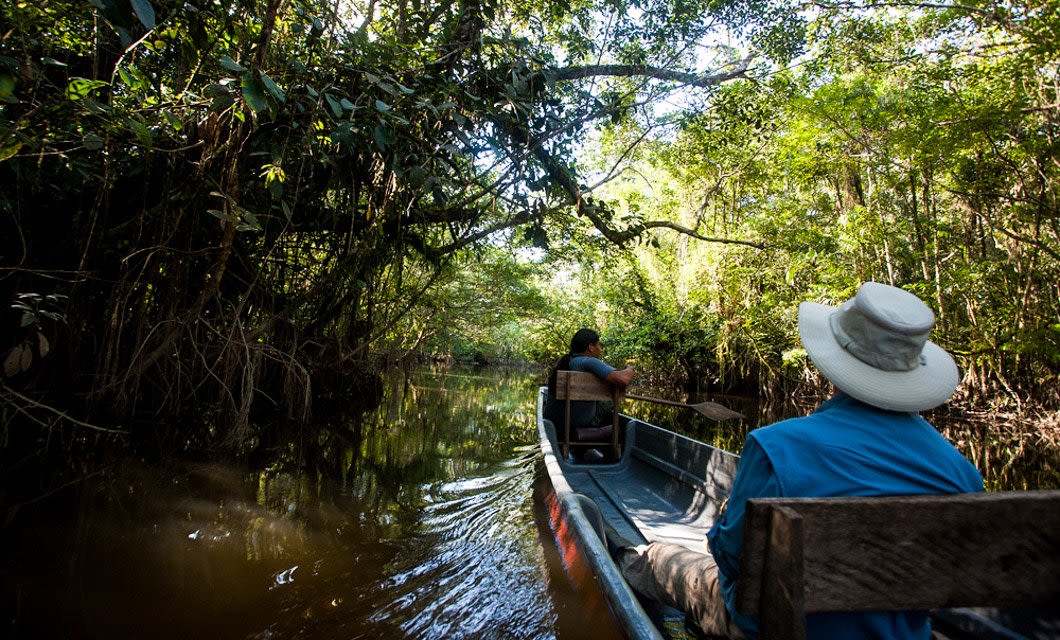 Two people ride in a wooden canoe along a narrow, tree-lined river in the lush Ecuadorian Rainforest. Sunlight filters through dense green foliage, casting shadows on the calm, brown water as their rainforest cruise winds quietly onward.