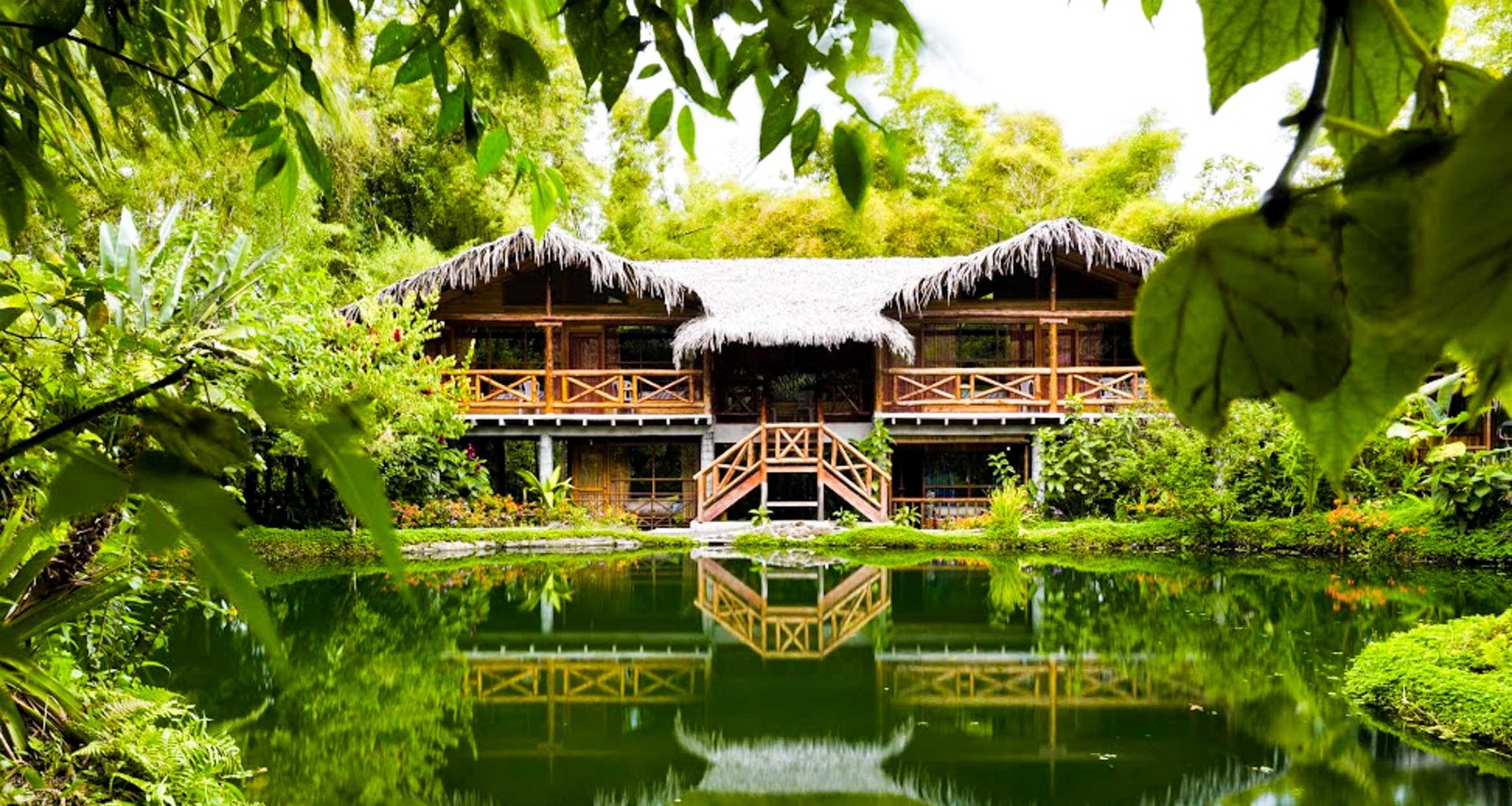 A rustic wooden lodge with a thatched roof is surrounded by lush greenery, reminiscent of the Amazon Rainforest, and reflected in a calm pond, framed by overhanging leaves and tropical plants.