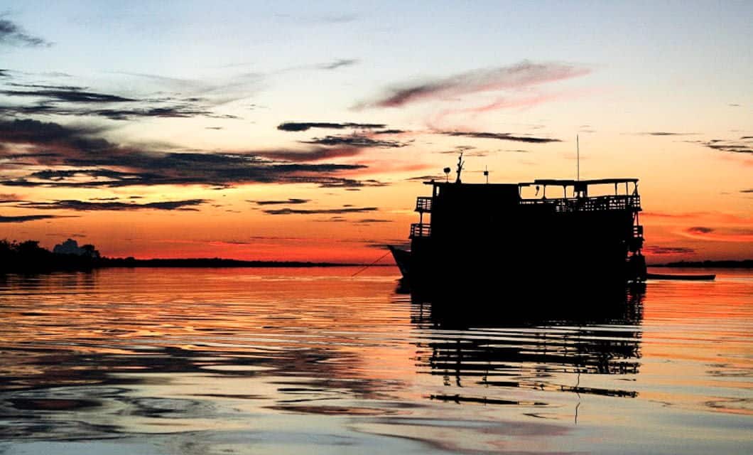 A large boat, the Tucano, is silhouetted against a vibrant orange and yellow sunset, floating on calm water with gentle ripples—an unforgettable scene from a Brazil Amazon River Cruise.