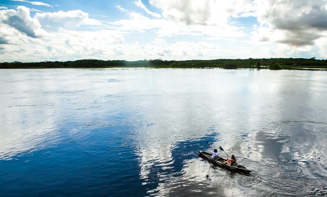 Two people paddle a small canoe on a calm, reflective river under a partly cloudy sky, with green trees lining the distant shore—an idyllic scene reminiscent of an Amazon cruise adventure.