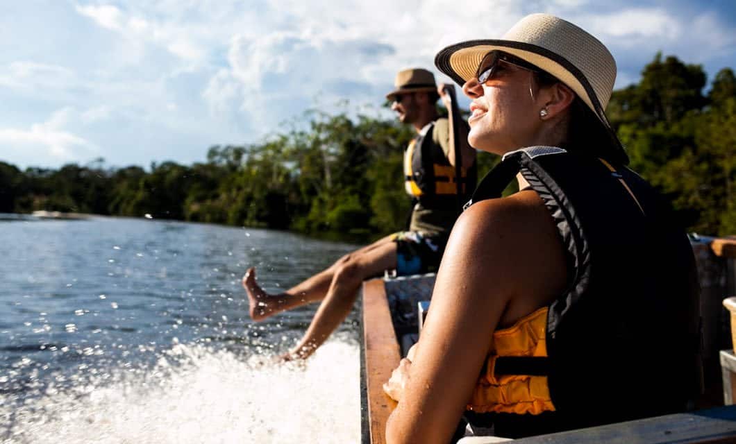 Two people wearing hats, sunglasses, and life jackets smile and relax on a boat during an Aria Amazon Cruise, with one dipping their feet in the water as the boat speeds along the Peruvian Amazon under a bright sky.