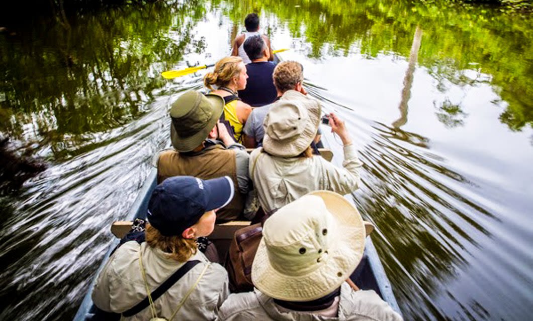 A group of people wearing hats and outdoor gear ride in a canoe along a calm, green river near Uakari Lodge, surrounded by lush vegetation, with some taking photos and others paddling.