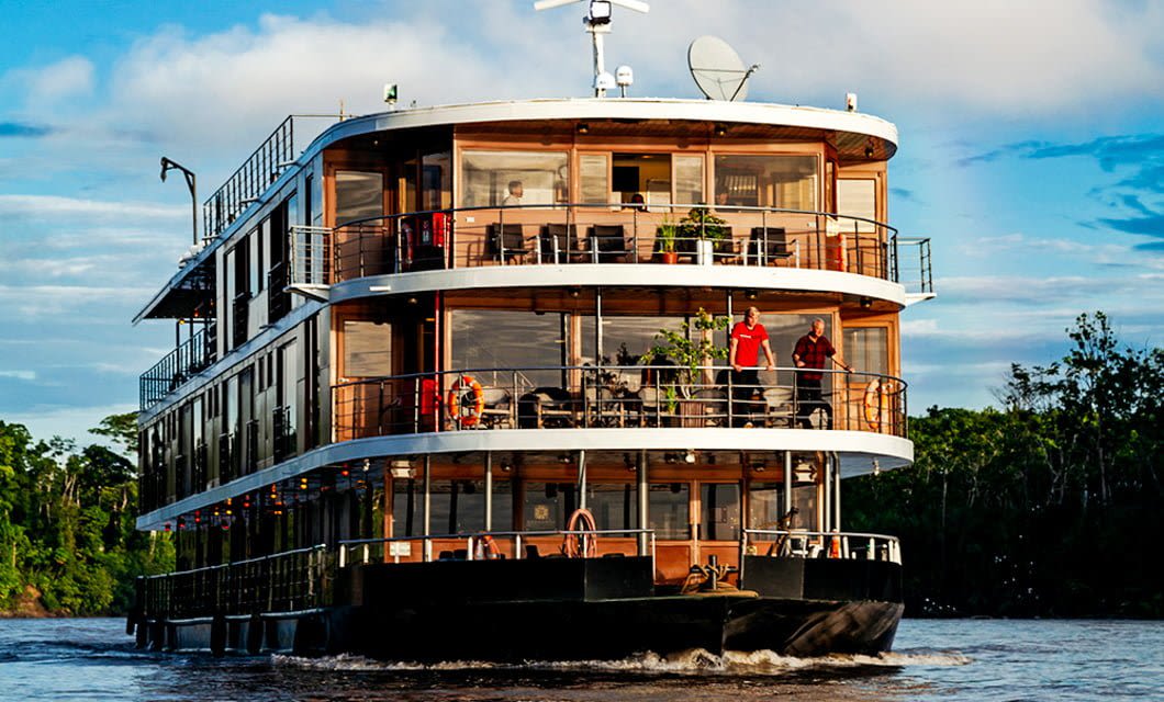 A modern, three-level Anakonda Amazon Cruises ship with large glass windows sails on the river, surrounded by lush greenery. People enjoy the scenery from the decks of this luxury Amazon cruise under a partly cloudy sky.