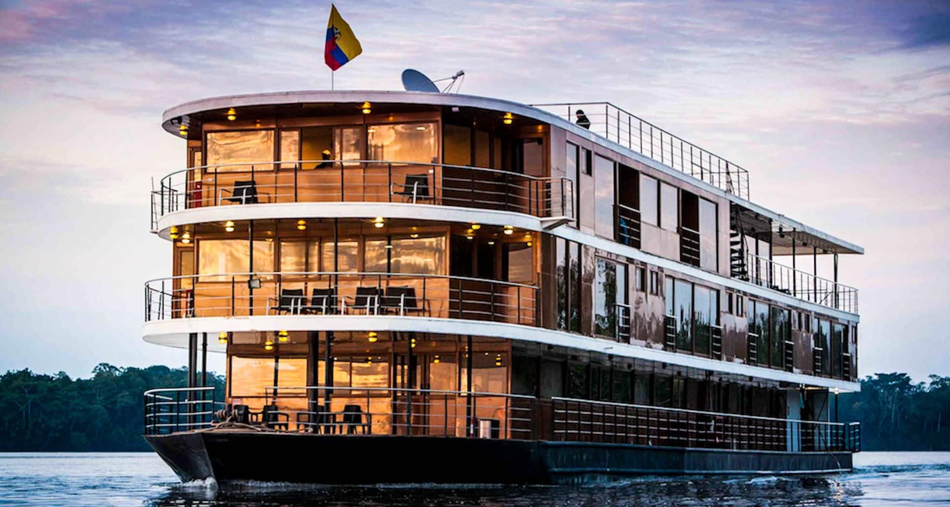 A modern, three-story Anakonda Amazon Cruises ship with large windows and balconies sails on calm water at sunset. Warm lights glow inside, an Ecuadorian flag flies atop the deck, and trees line the distant shore.