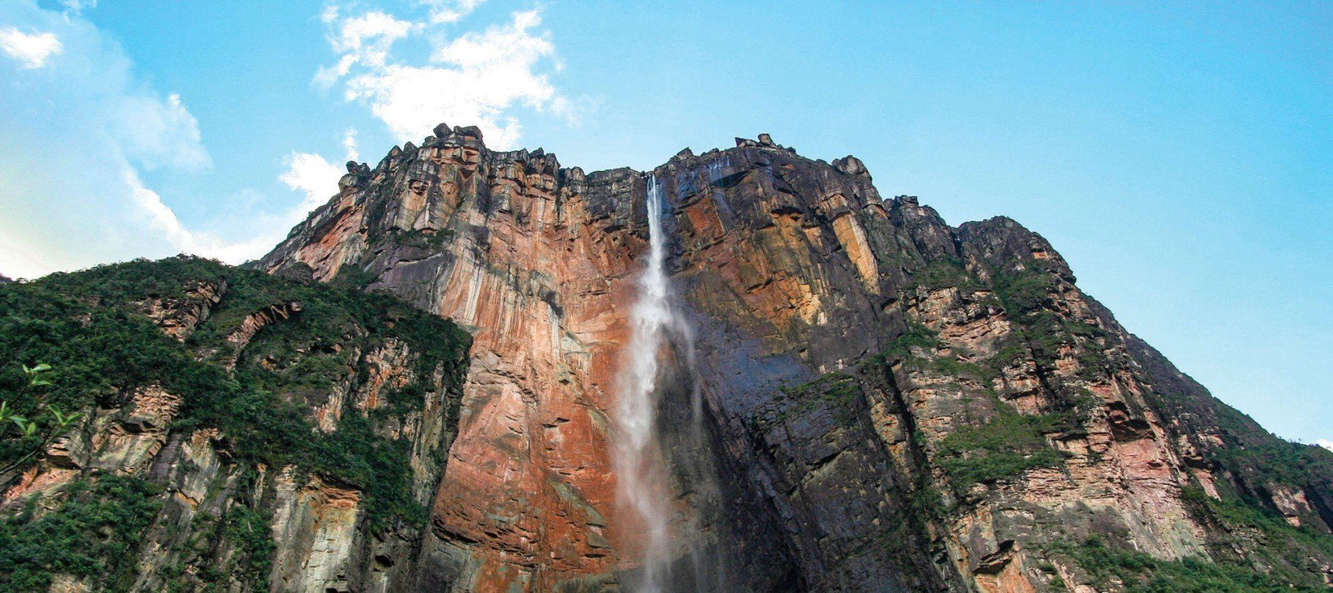 A tall, narrow waterfall cascades down a steep, reddish-brown cliff surrounded by lush vegetation and a bright blue sky—an iconic sight on many Venezuela tours and perfect for unforgettable Venezuela vacations.