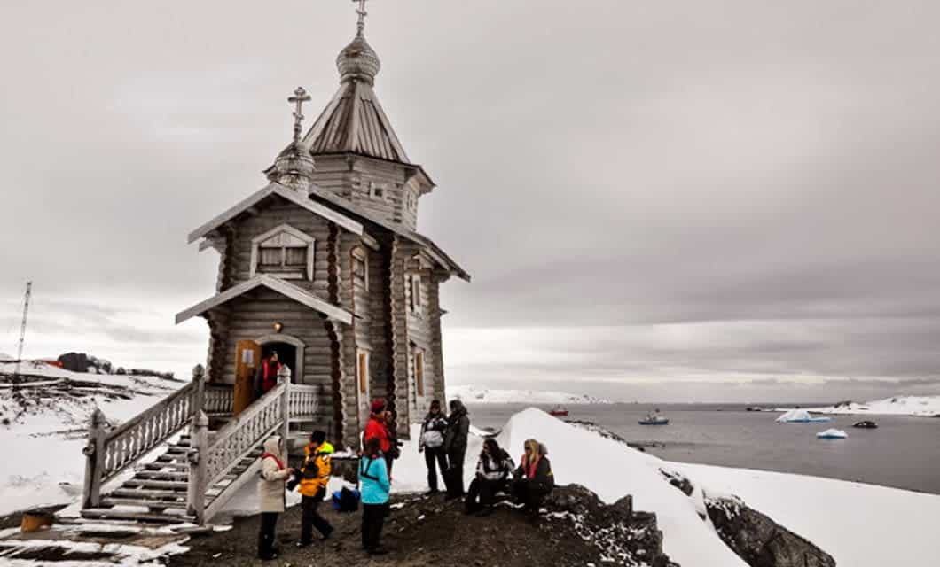 A small wooden church with a steeple stands on a snowy landscape near the water, as bundled-up people from an Antarctic expedition gather outside. Icebergs float in the water under a cloudy sky.