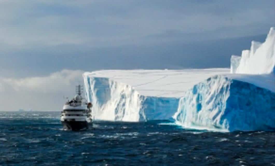 A large yacht sails near a massive, flat-topped iceberg in a dark blue, choppy sea under a cloudy sky, evoking the spirit of an Antarctica Cruise from Ushuaia. The iceberg has steep, icy blue sides and a snow-covered top.
