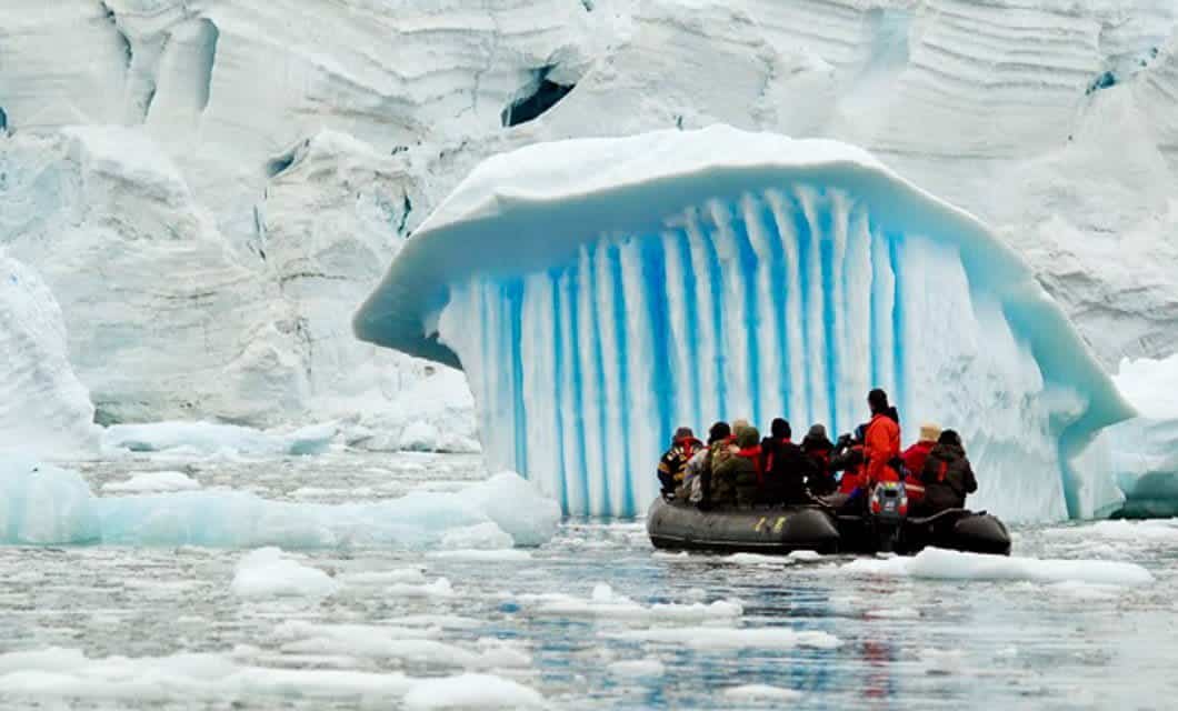 A group of people wearing cold-weather gear sits in a rubber boat on icy water near a glacier, observing a snow-covered ice formation with striking vertical blue stripes during an unforgettable Antarctica cruise.