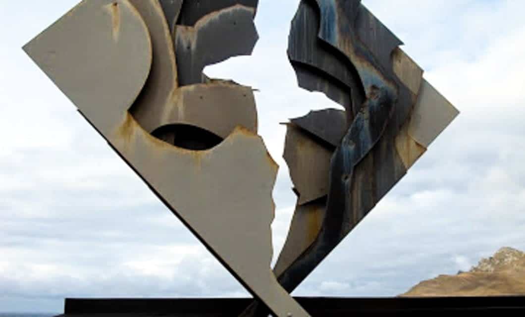 Abstract metal sculpture with jagged, torn edges stands outdoors against a cloudy sky and distant hills, reminiscent of forms seen on an Antarctica cruise. Intersecting shapes evoke the raw beauty of an Antarctic expedition.