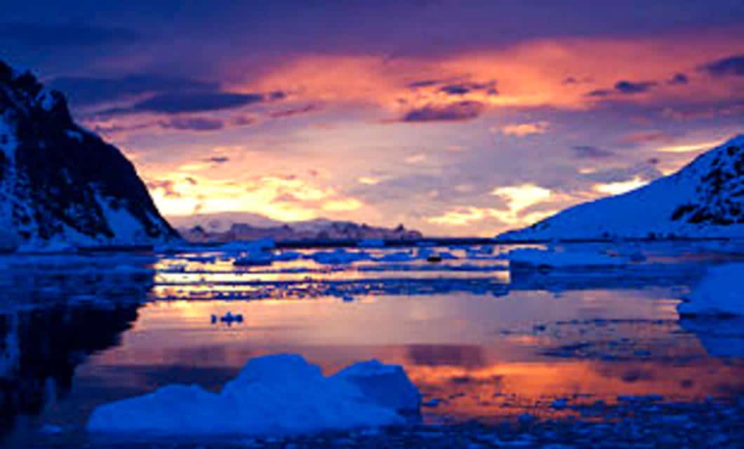 A dramatic sunset over icy waters with floating icebergs, reflecting vibrant orange, pink, and purple hues in the sky; snow-covered mountains on both sides set the scene for a breathtaking Antarctic expedition.