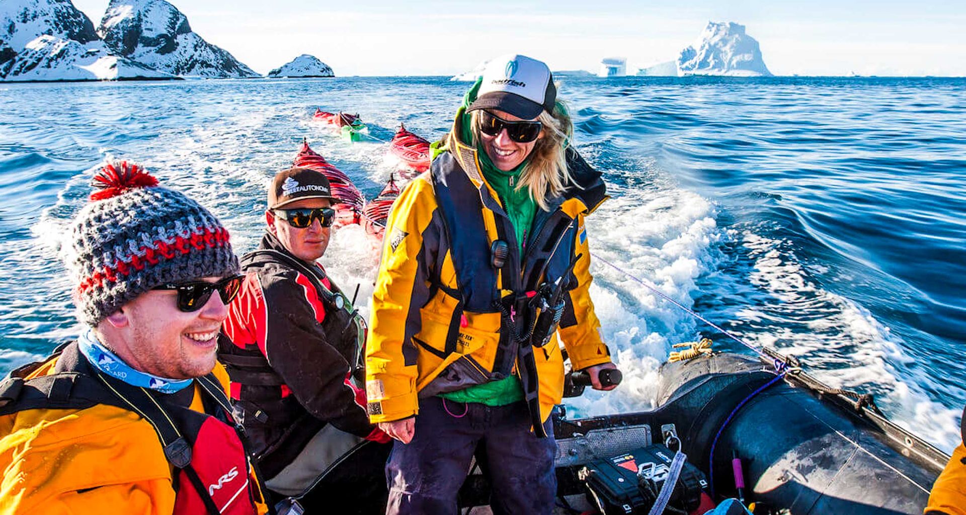 Three people in warm jackets and hats ride a small inflatable boat through icy blue water, snow-covered mountains, and floating icebergs—a snapshot from an unforgettable Antarctic expedition under a clear sky.