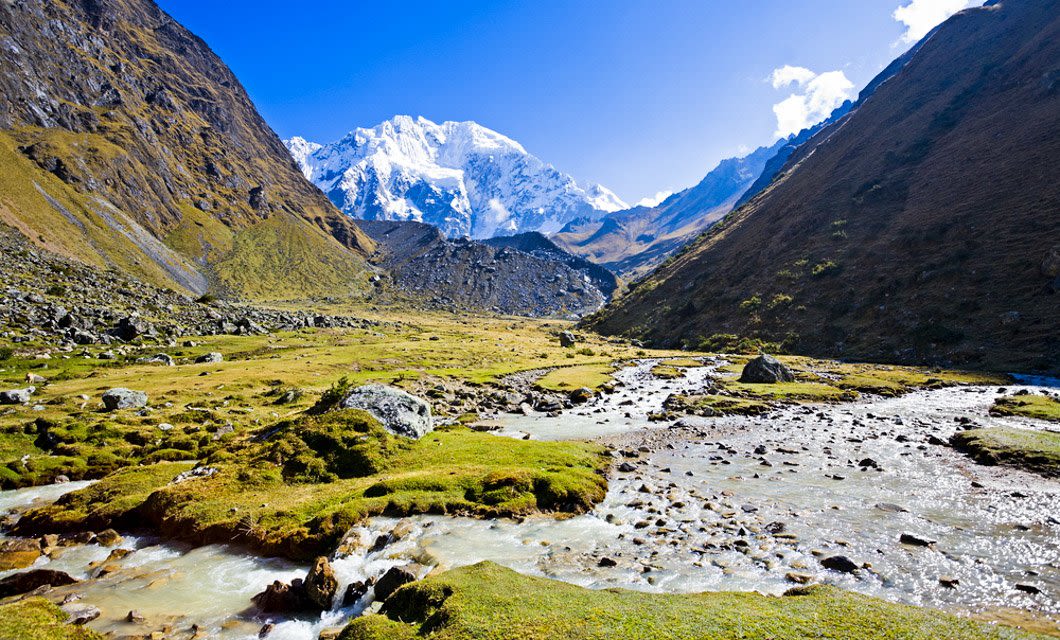 A clear stream winds through a grassy valley surrounded by steep mountains, with snow-capped peaks in the distance—an iconic scene along the Salkantay Trek to Machu Picchu under a bright blue sky.