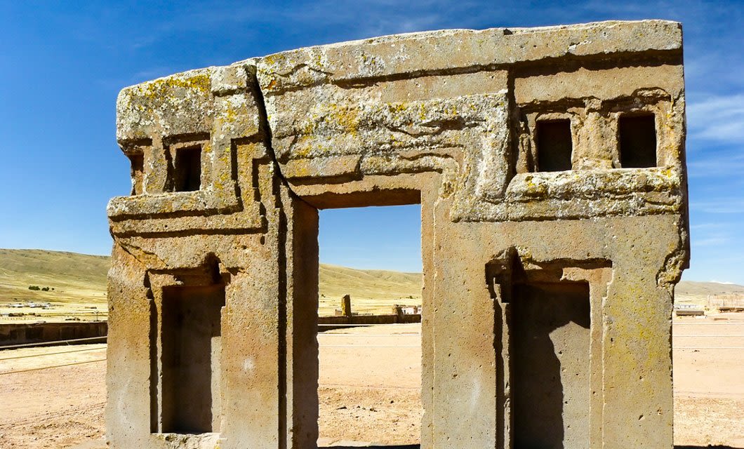 Ancient stone gateway with geometric carvings and rectangular openings, set in a dry, open landscape under a blue sky with distant hills—a silent witness to the Travels of the Incas near La Paz.