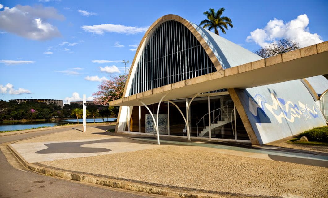 A modernist church with a curved blue and white roof, large windows, and patterned sidewalk sits beside a lake in one of Brazil's historic cities, framed by palm trees and blue sky in the background.