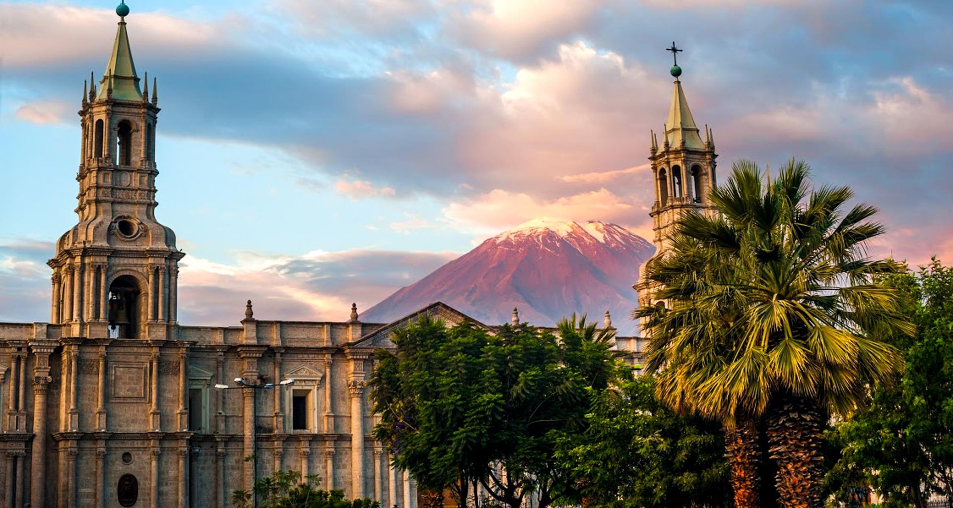 A historic stone cathedral with two towers stands before lush trees and palm trees, embodying Peruvian culture, as a snow-capped volcano rises in the background under a colorful sunset sky—an unforgettable travel scene in Peru.