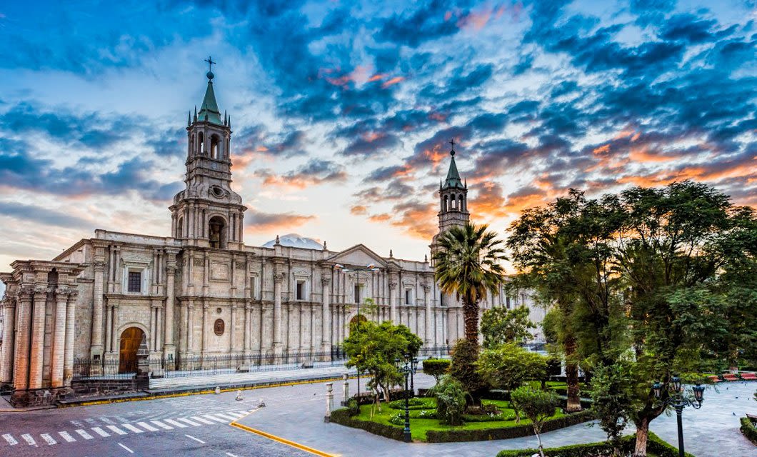 A grand stone cathedral with two towers stands beside a tree-lined park, embodying the rich culture of Peru, under a vibrant sunset sky with scattered clouds—a dream scene for travel enthusiasts.