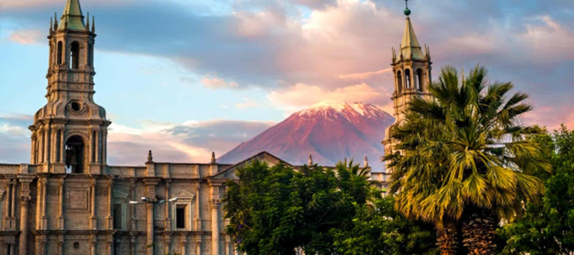 Two ornate church towers rise above leafy trees, with a snow-capped volcano in the background under a colorful sunset sky—an iconic scene often featured in Arequipa Tours.
