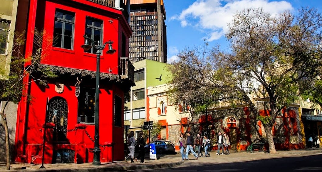 A brightly colored red building stands on a city street corner in Argentina, with people walking by on the sidewalk. Leafless trees line the street and other colorful buildings are visible under a blue sky with clouds.