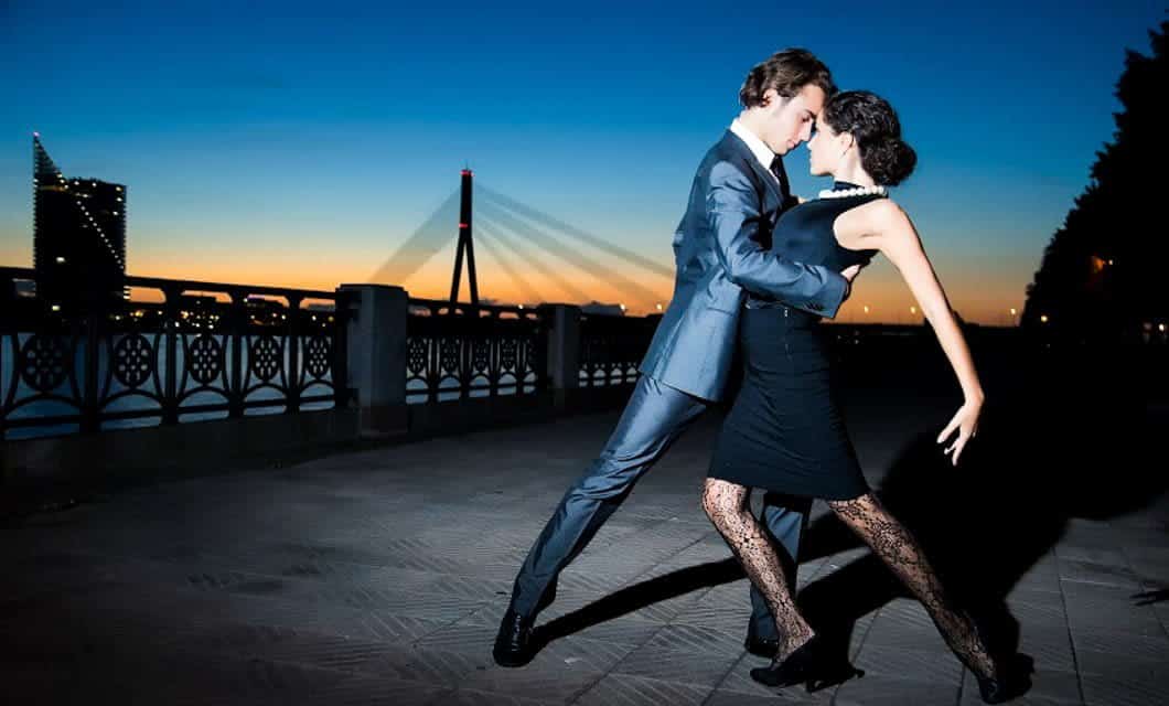 A man in a suit and a woman in a black dress and patterned tights perform a tango pose outdoors at dusk, evoking the passion of South America, with a bridge and cityscape in the background.