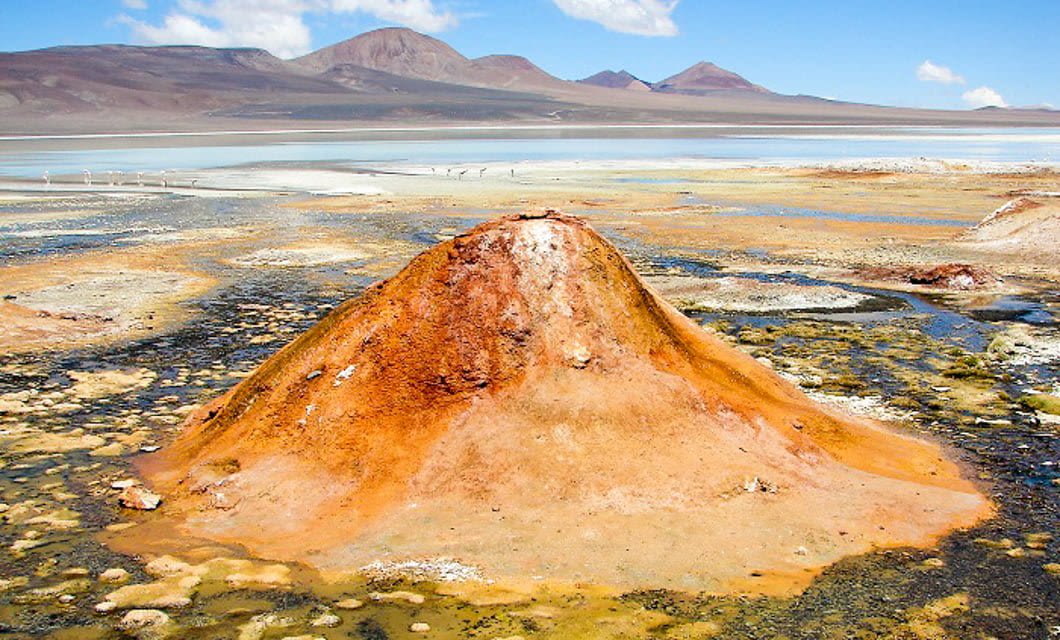 A rusty orange, cone-shaped geothermal mound sits in a colorful, mineral-rich landscape with patches of water, under a blue sky and the distant Andes mountains near Velasco.