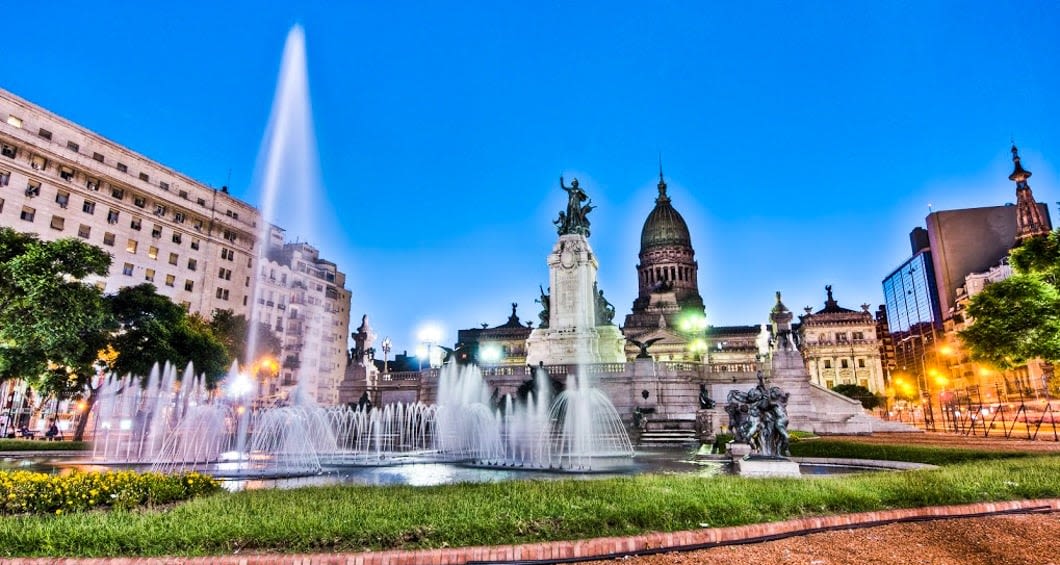 A large ornate fountain with statues, reminiscent of waterfalls, stands before a grand domed building, surrounded by historic architecture and trees, all illuminated against a clear evening sky.