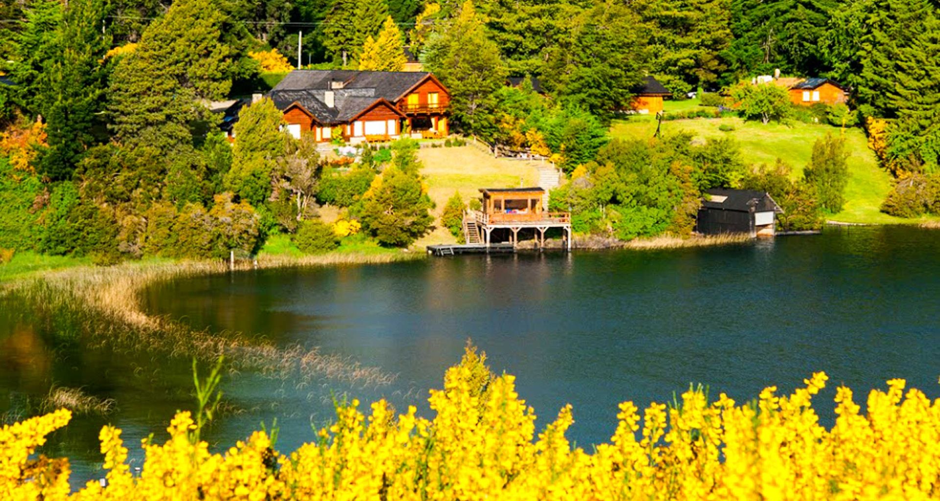Wooden cabins surrounded by green trees and a lush lawn sit on a hillside overlooking a calm lake, with vibrant yellow flowers in the foreground. Enjoy a world class Alpine experience as a small dock extends into the water.