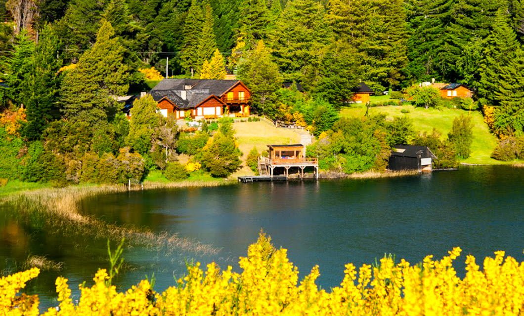 A wooden house surrounded by lush green trees sits beside a calm lake, echoing the natural beauty of Argentina. Bright yellow flowers bloom in the foreground under a sunny sky, capturing the best of Argentina's serene landscapes.